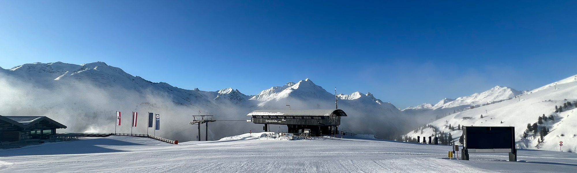 Panoramaaufnahme eines verschneiten Skigebiets mit Berghütten, Liften und umliegenden Alpengipfeln bei klarem, blauem Himmel.