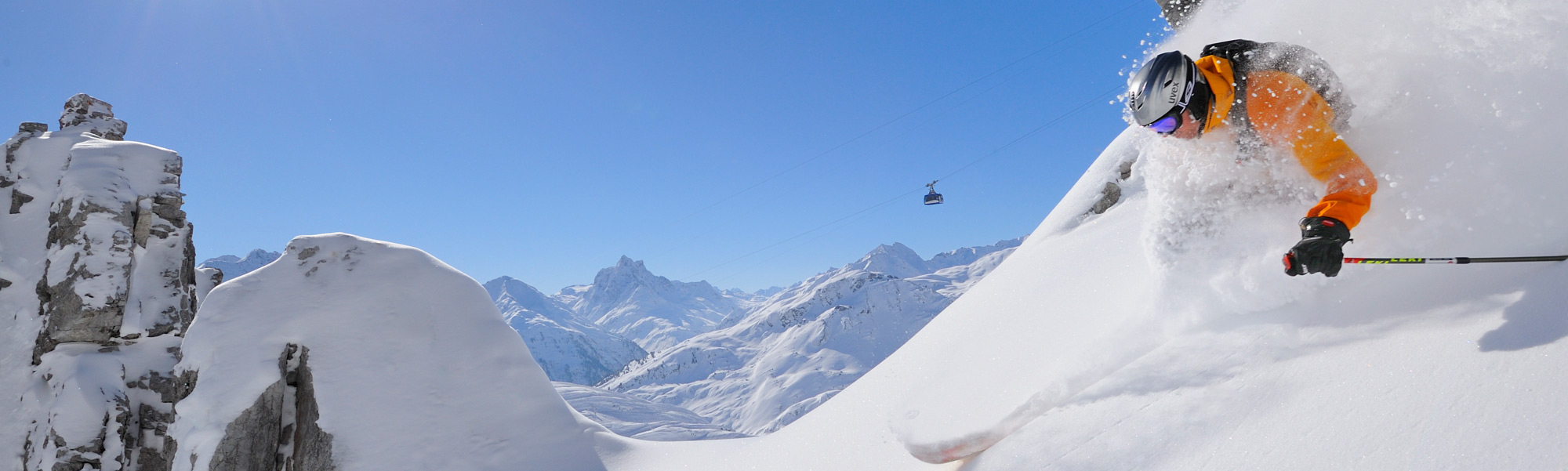 Skifahrer in orangefarbener Jacke fährt dynamisch durch tiefen Pulverschnee.