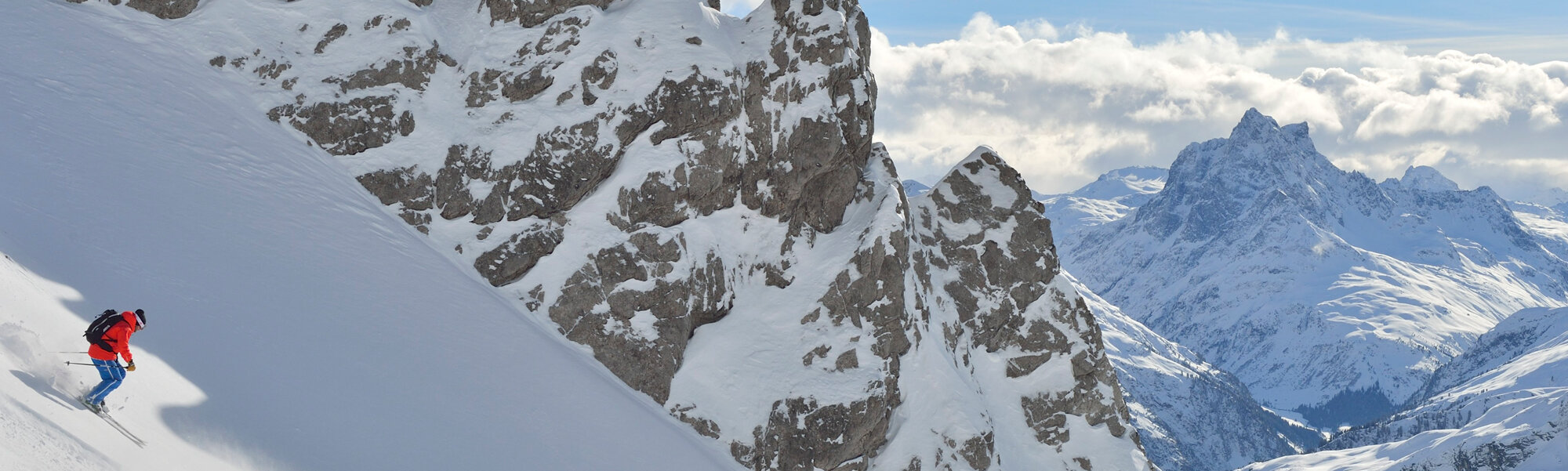 Skifahrer fährt entlang steiler Schneeflanke, felsige Bergwand im Hintergrund.