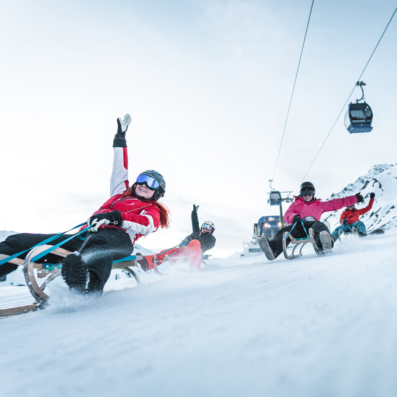 Gruppe fröhlicher Rodler auf präparierter Piste mit Gondelbahn und Winterlandschaft im Hintergrund.